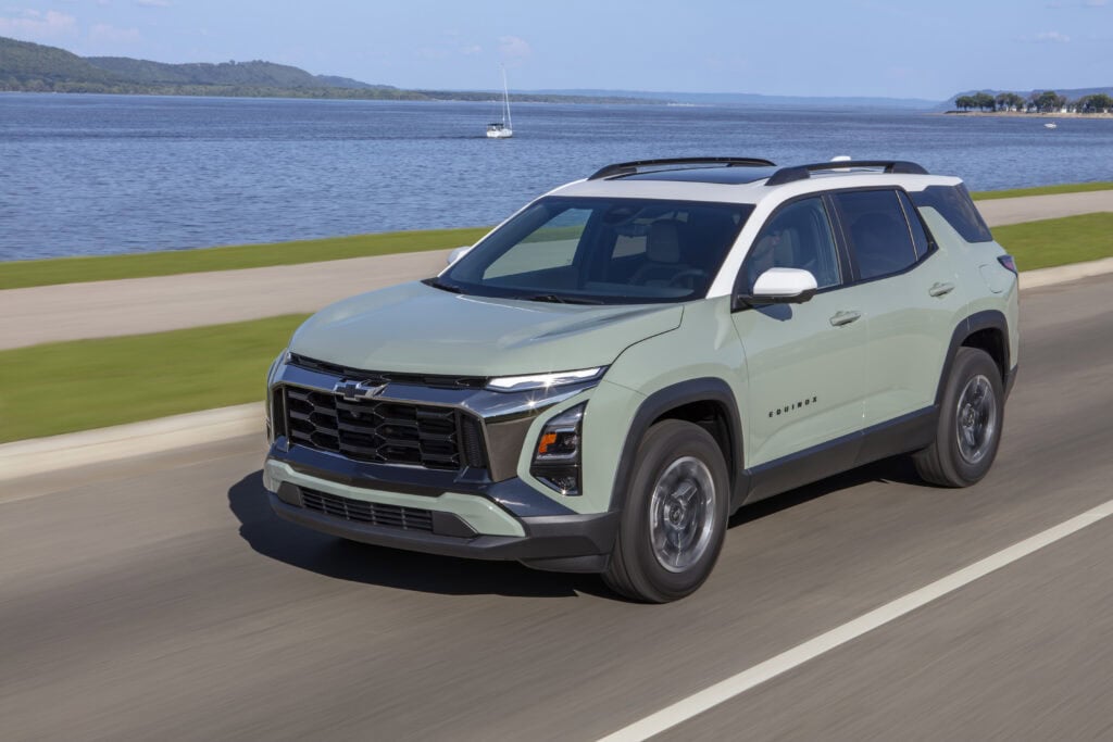 Front 3/4 view of a 2025 Chevrolet Equinox ACTIV in Cacti Green with Summit White roof driving near a large body of water.