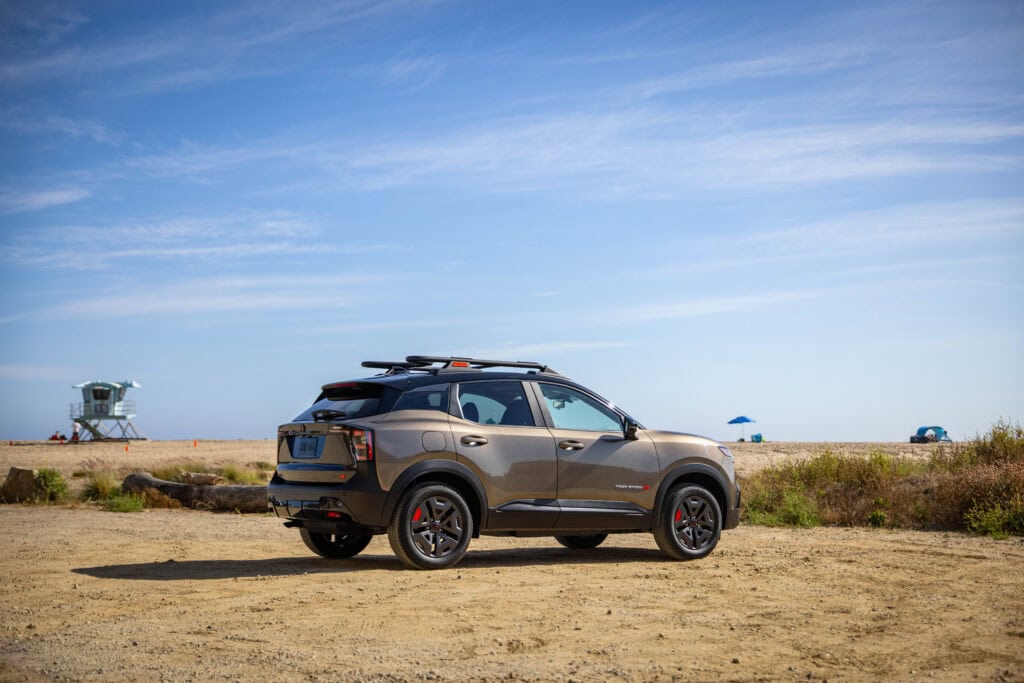 Angled rear view of the 2027 Nissan Kicks Rock Creek parked on a beach, beach tents can be seen behind.