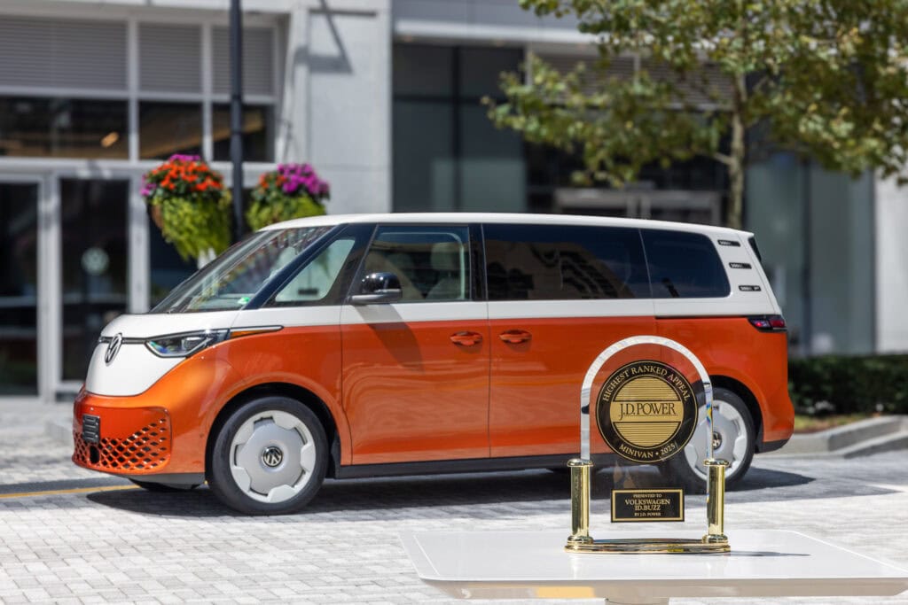 The 2026 VW ID Buzz electric bus sitting in a courtyard with a building behind it that has flowers hanging from hanging baskets and in the foreground, the JD power award