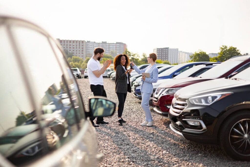 A car salesperson hands the keys to a new car owner as their partner claps in the background. They are at a car dealership.