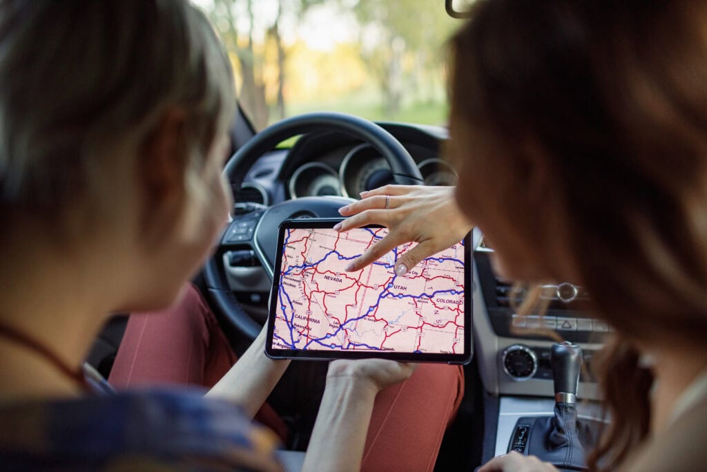 Happy young two women driver tourist travelers with digital tablet USA American map at rental car vehicle