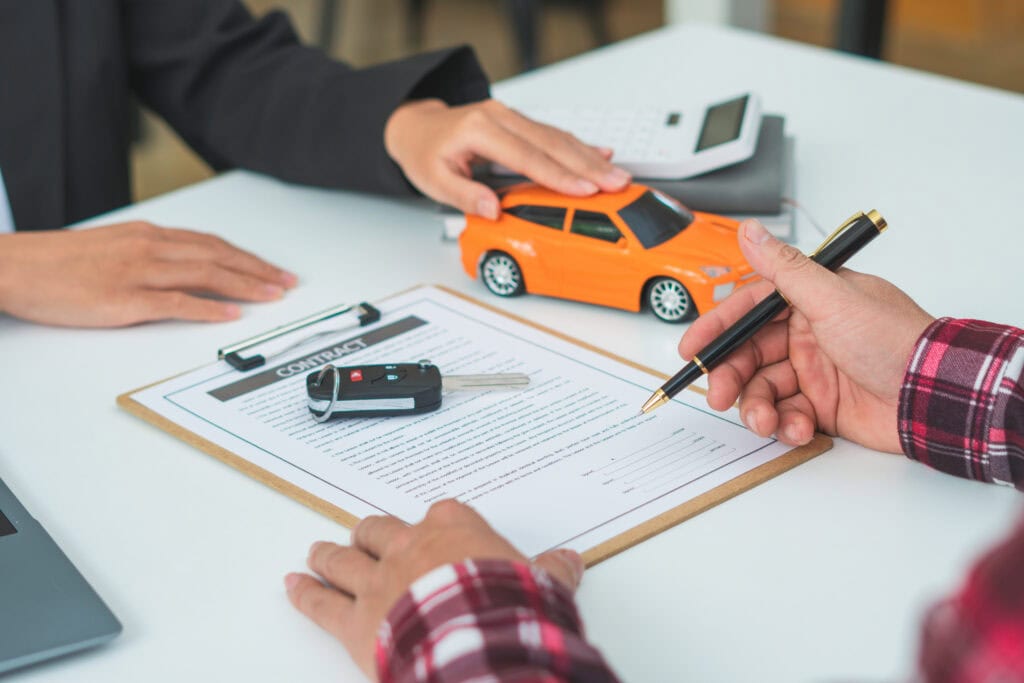 A man signing a car document or a lease paper. Writing signature on contract or agreement. Buying or selling new or used vehicle. Car keys on table. Warranty or guarantee. Customer or salesman.