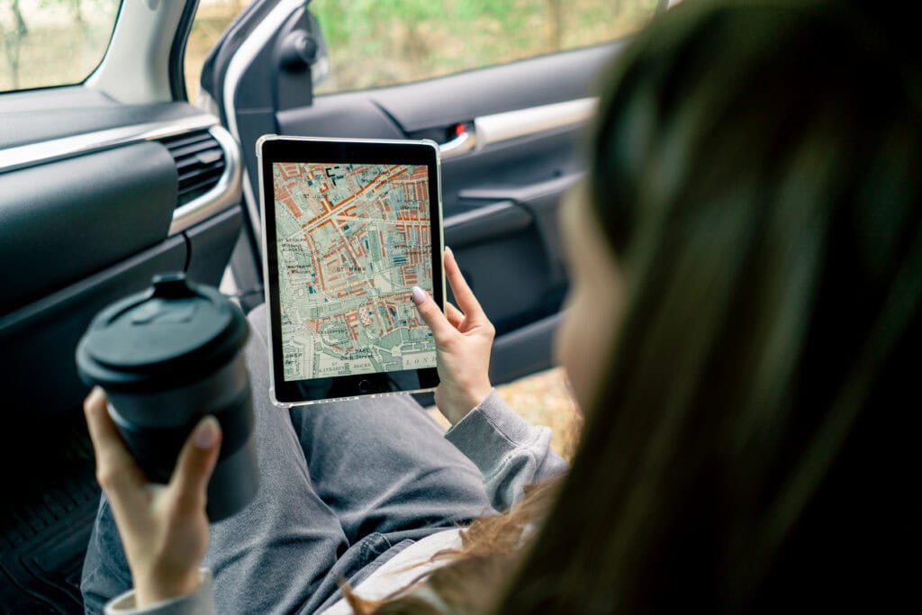 A girl sits in a car in the front passenger seat with a cup of tea and plots a route through the forest using a map on a tablet